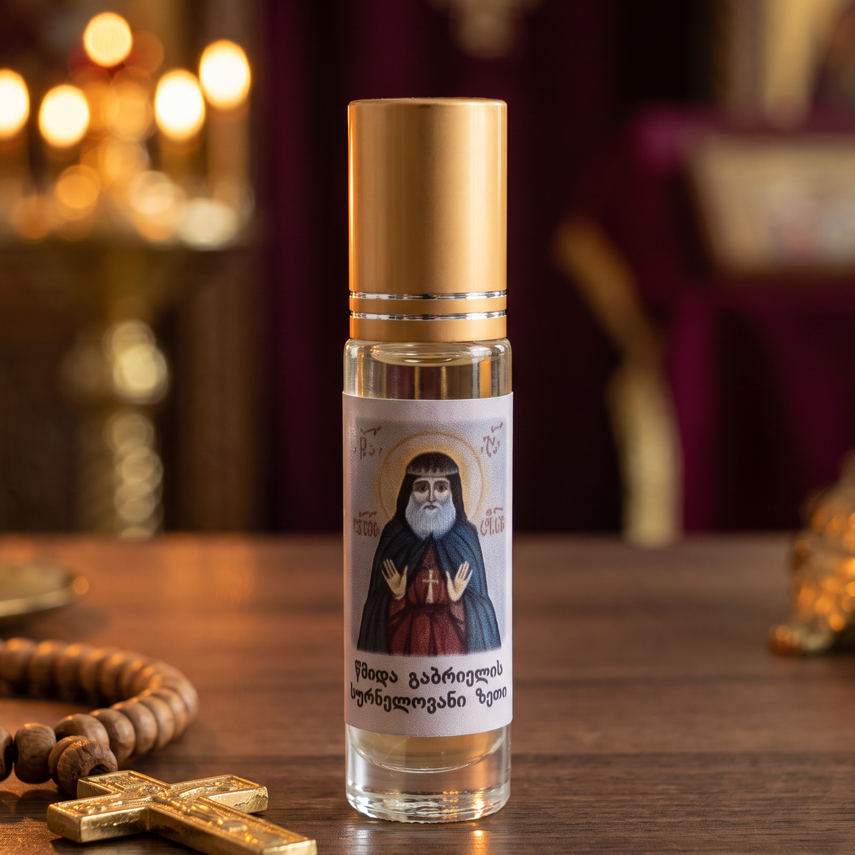 A small clear glass bottle with a gold cap stands on a wooden table, labeled with an icon of a religious figure. A wooden rosary and a gold cross rest beside it, with warm blurred candles and ornate decor in the background.