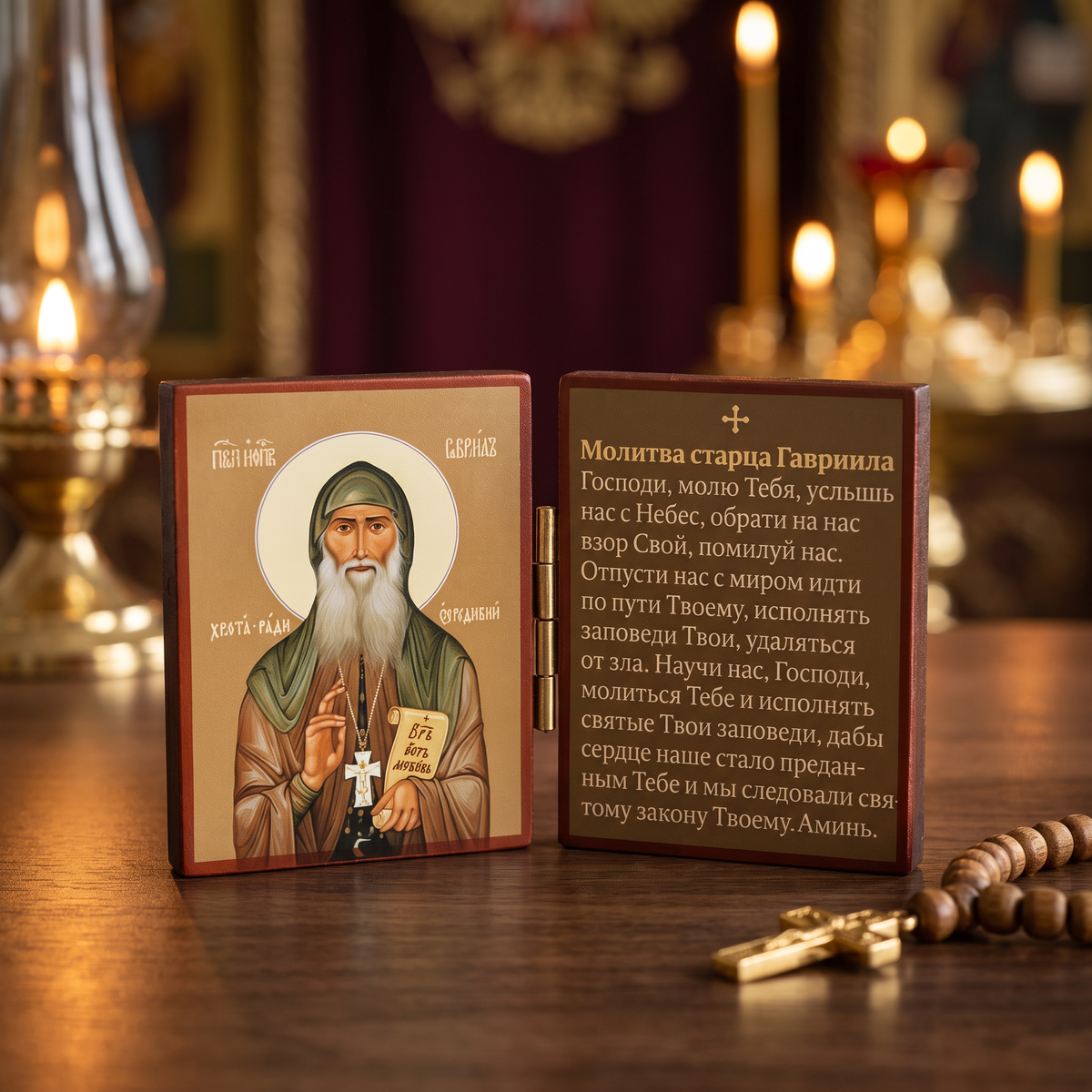 A wooden Orthodox prayer box shown open on a wooden table, with a painted icon of Saint Gavrilius on the left panel and a printed prayer text on the right panel. A rosary lies nearby on the table, and blurred candles and warm interior decor appear in the background.